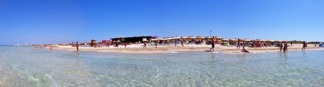 Spiaggia affollata con ombrelloni colorati e persone in acqua sotto un cielo blu.