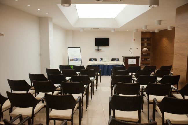 Sala conferenze con sedie nere, un podio e uno schermo televisivo.