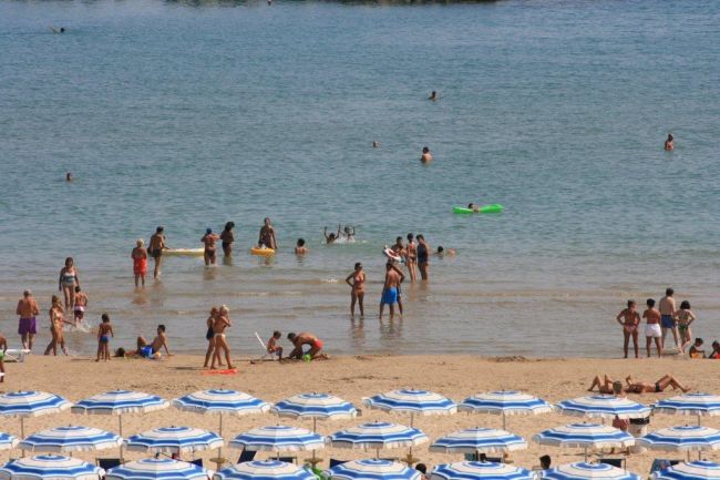 Persone in acqua poco profonda vicino alla riva, ombrelloni bianchi e blu sulla spiaggia sabbiosa.