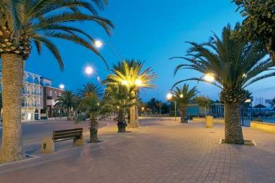 Viale pedonale di sera con palme e lampioni accesi.