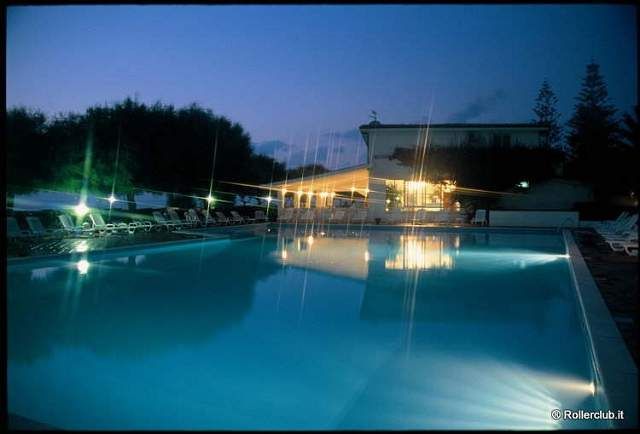 Piscina illuminata di sera con sedie a sdraio attorno e un edificio sullo sfondo.
