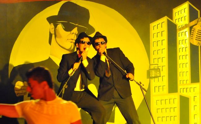 Due uomini in abiti neri e occhiali da sole cantano su un palco con un murale dietro.