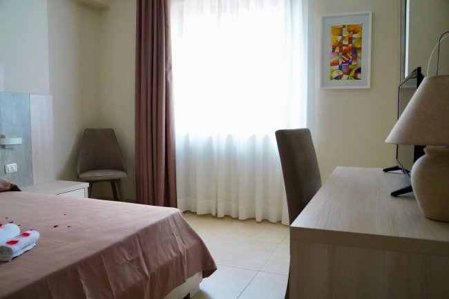 Camera con letto matrimoniale, tendaggi chiari e scrivania.