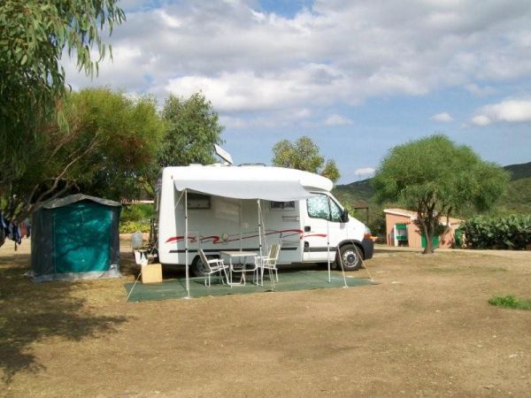 Camper bianco parcheggiato, con tenda e sedie all'aperto, alberi sullo sfondo.