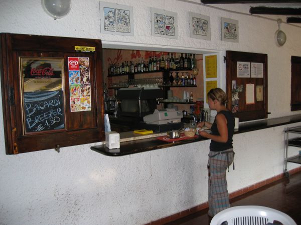 Donna prepara bevande al bancone di un bar con mensole di bottiglie dietro.