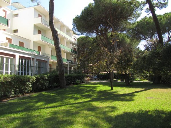 Edificio bianco con balconi verdi, alberi e prato curato.