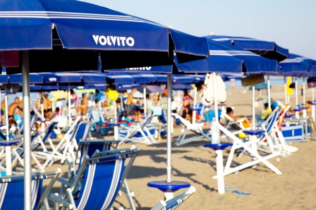 Una spiaggia con sedie a sdraio e ombrelloni blu; persone in background.