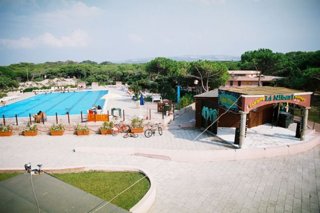 Piscina olimpionica all'aperto, bar e biciclette parcheggiate, circondata da verde.