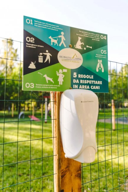 Cartello con regole per l'area cani e un dispenser su un palo in un parco verde.