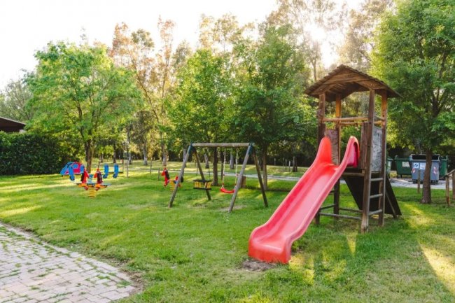 Area giochi all'aperto con scivolo rosso, altalene e alberi circostanti.