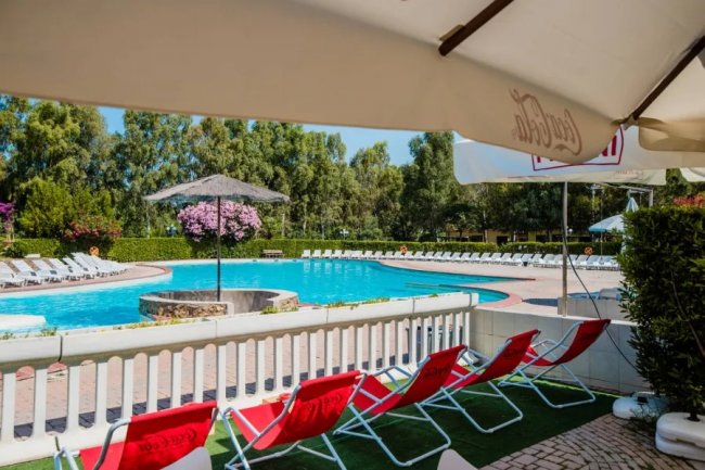 Piscina all'aperto con sedie a sdraio rosse, ombrelloni e alberi sullo sfondo.