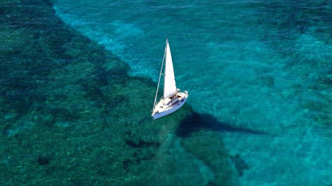 Una barca a vela bianca naviga su un mare cristallino visto dall'alto.