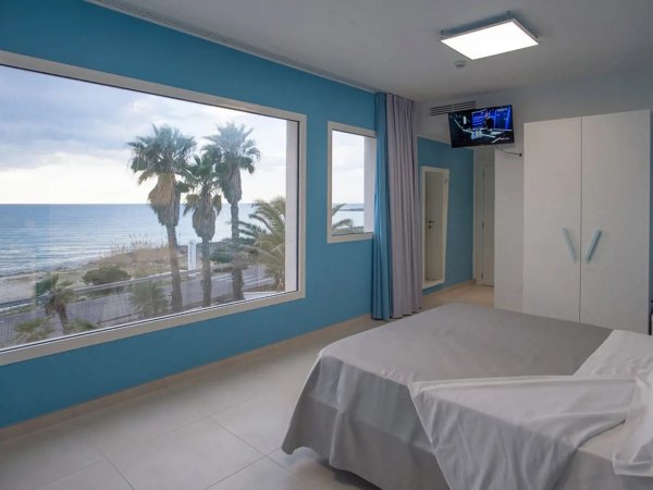 Camera con vista mare, letto bianco, pareti azzurre e grande finestra.