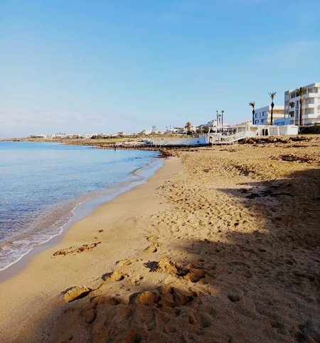 Spiaggia sabbiosa con mare calmo e edifici bianchi sullo sfondo sotto un cielo azzurro.