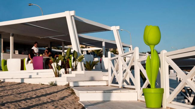 Terrazza di un bar sulla spiaggia con arredi bianchi e un grande vaso verde a forma di tulipano.