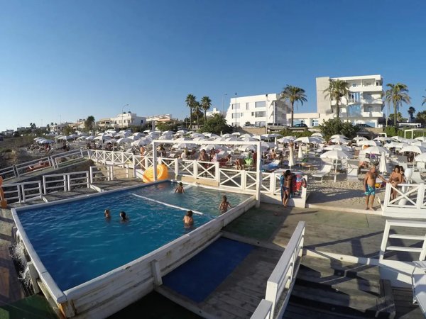 Una piscina con persone che nuotano, ombrelloni bianchi e un edificio sullo sfondo.