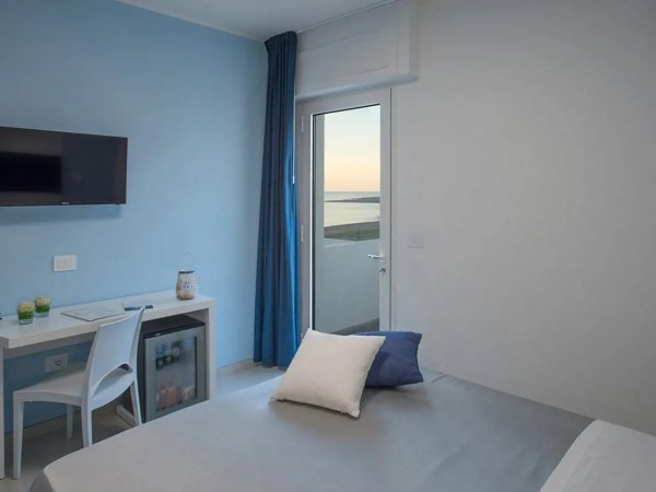 Camera moderna con pareti blu, balcone con vista mare, scrivania, TV e letto con cuscini.