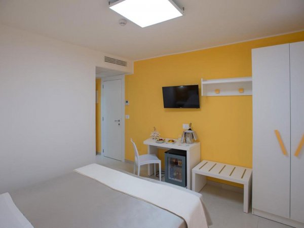 Camera luminosa con pareti gialle, TV, scrivania, minibar, armadio e letto con coperta chiara.