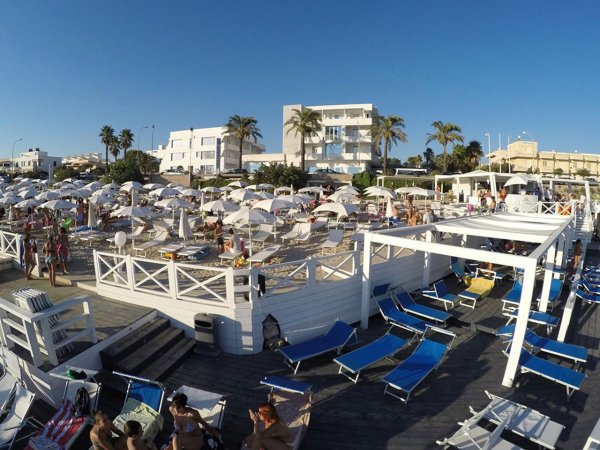 Spiaggia affollata con lettini blu, ombrelloni bianchi, e edifici bianchi sullo sfondo.