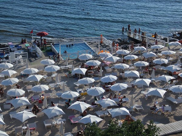 Spiaggia affollata con ombrelloni bianchi, sedie a sdraio, piscina e mare.