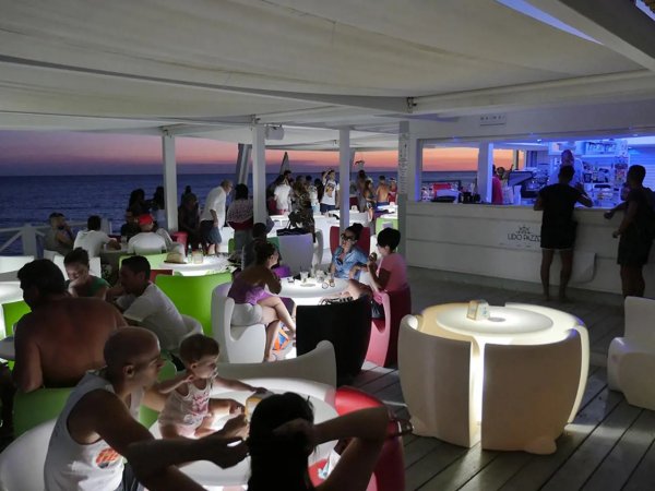 Lounge bar in spiaggia con tavoli luminosi e persone sedute al tramonto.