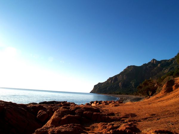 Costa rocciosa con terra rossa, mare calmo e montagne in lontananza sotto un cielo azzurro.