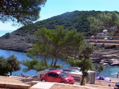 Litorale con auto parcheggiata, alberi e campeggio; collina verdeggiante sullo sfondo.