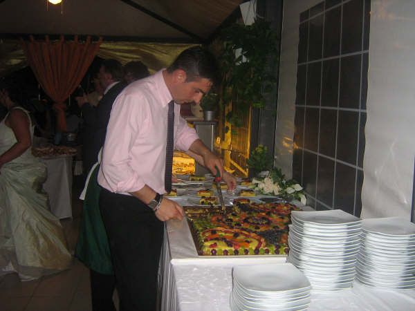 Uomo in camicia rosa che taglia una grande torta alla frutta su un tavolo da buffet.